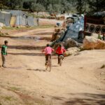 Children ride bicycles on a dirt road near makeshift tents in Idlib, Syria, capturing innocence and childhood joy.