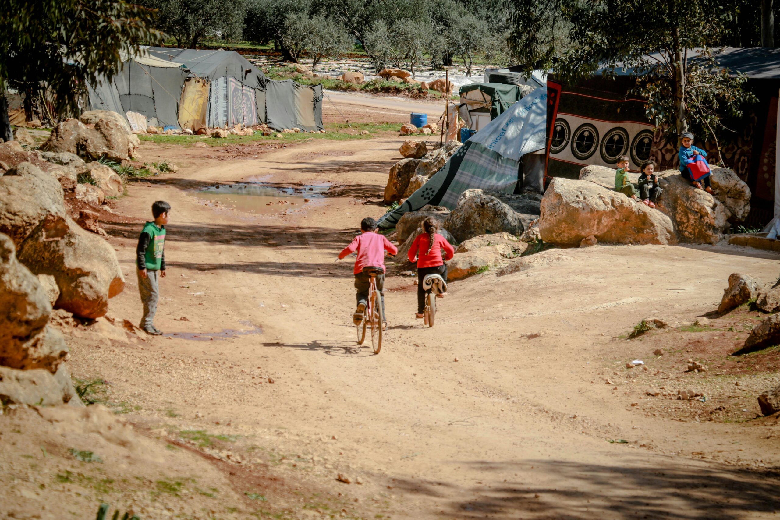 Children ride bicycles on a dirt road near makeshift tents in Idlib, Syria, capturing innocence and childhood joy.